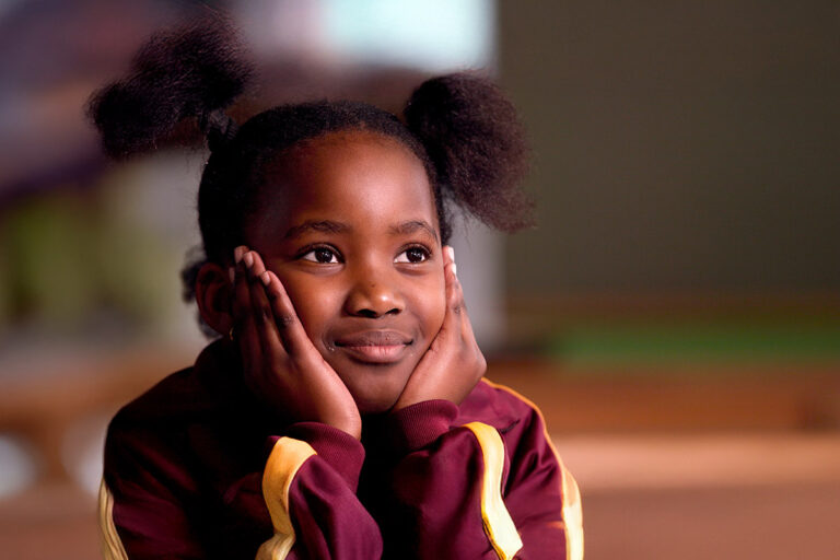 slider-spenden-2025 Afrikanisches Schulmädchen stützt ihr Gesicht in ihre Hände und lächelt